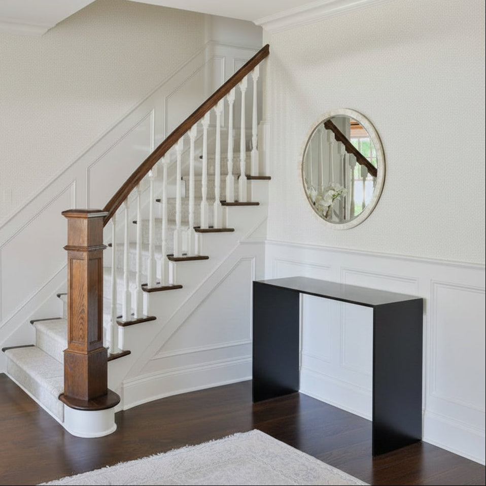 An elegant interior entryway featuring a staircase with a dark wood handrail and white spindles, a light blue console table with decorative items, a round wall mirror, and a textured orange starburst ceiling light.