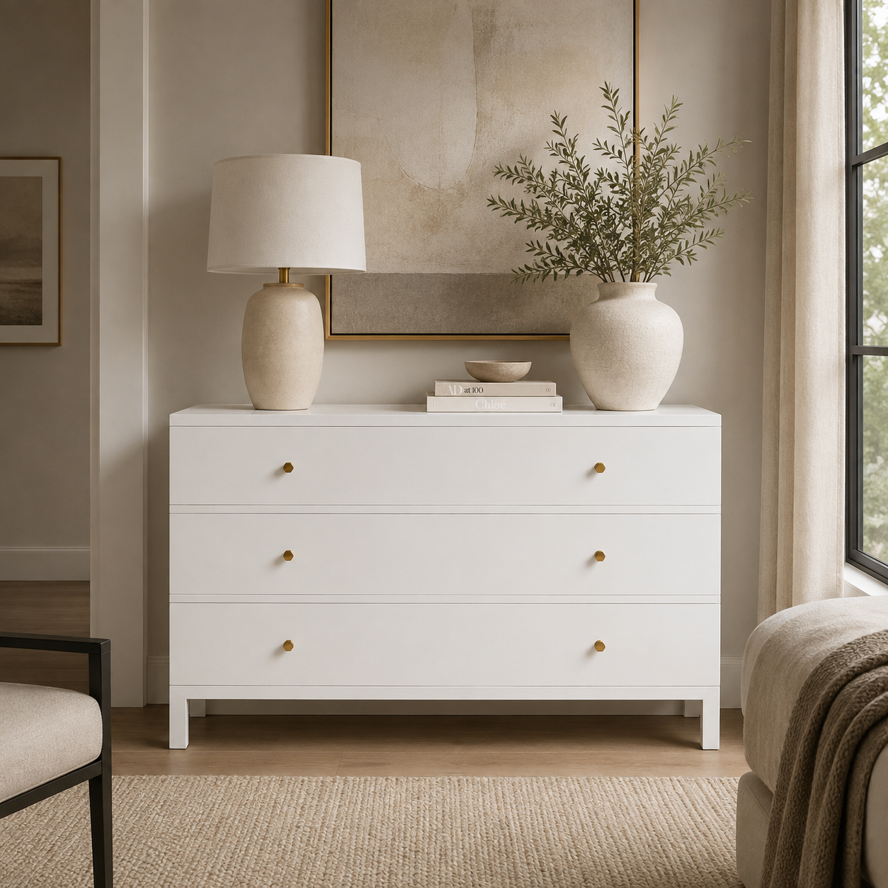 A white three-drawer dresser in a styled bedroom setting, topped with a cream table lamp and a large ceramic vase holding green branches, with small books and a bowl on top.