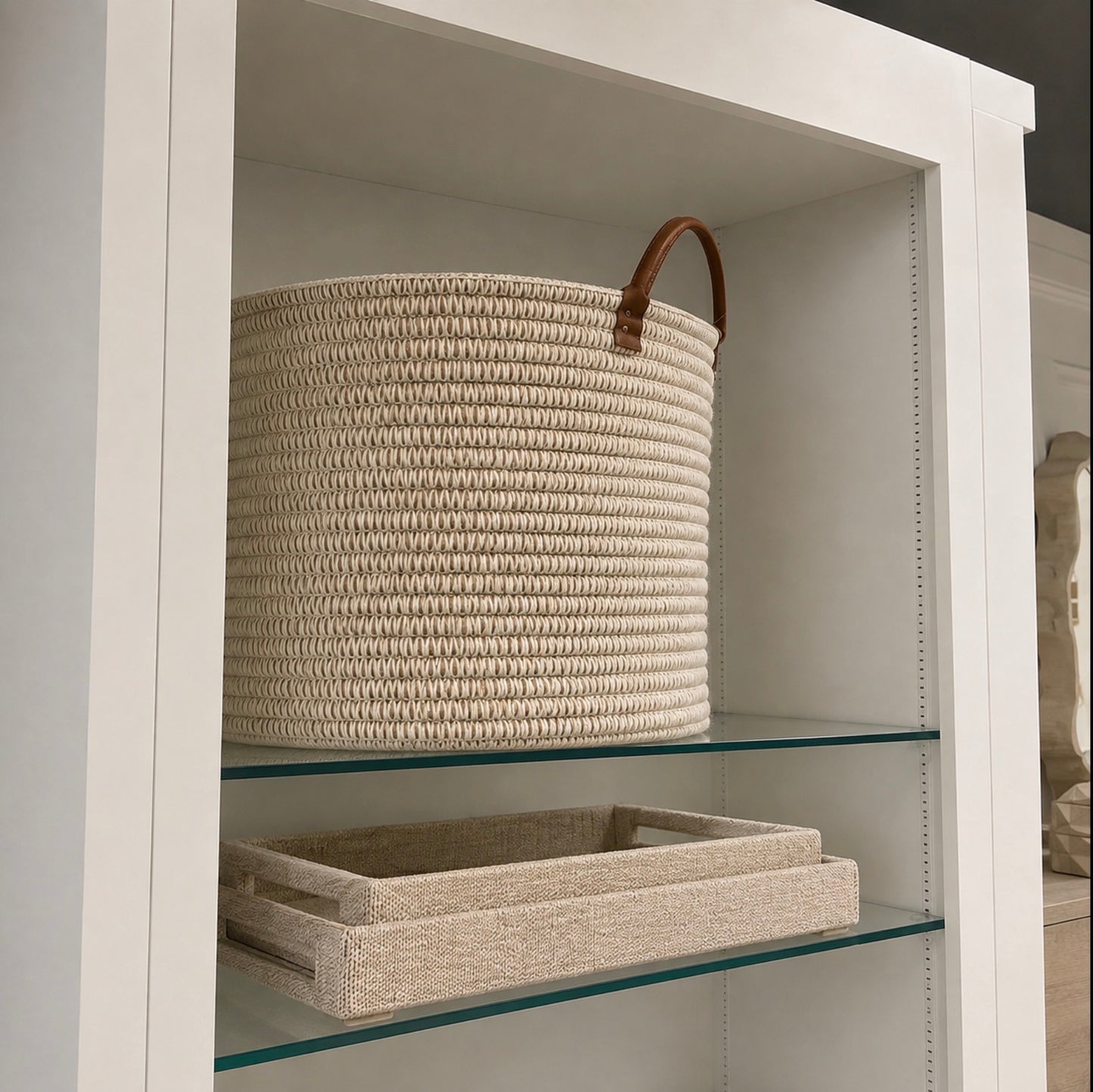 A large cylindrical woven storage basket in natural beige rope with a brown leather handle, displayed on a glass shelf inside a white cabinet. A matching woven tray sits on the lower shelf.