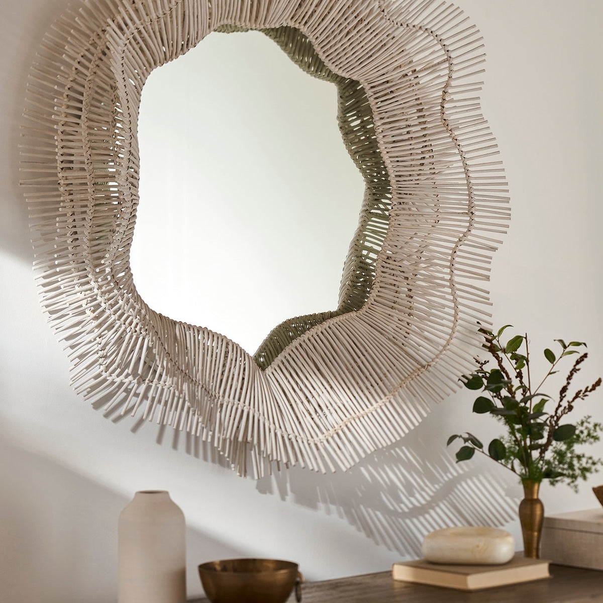 An organic-shaped wall mirror with a wide, sculptural frame made of many thin, light-colored sticks radiating outward, mounted on a beige wall above a styled tabletop with a vase, bowl, and small plant.