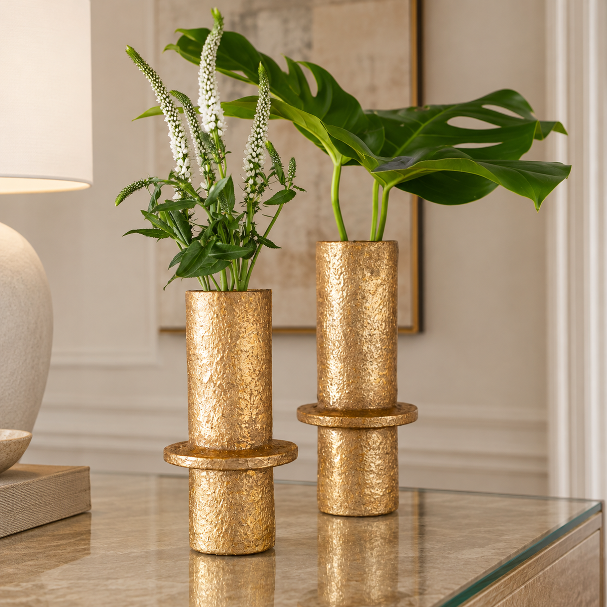 Two stacked gold metallic cylindrical vases with a textured finish, one holding white flowers and the other a green monstera plant, displayed on a glass-topped table in a styled interior setting.