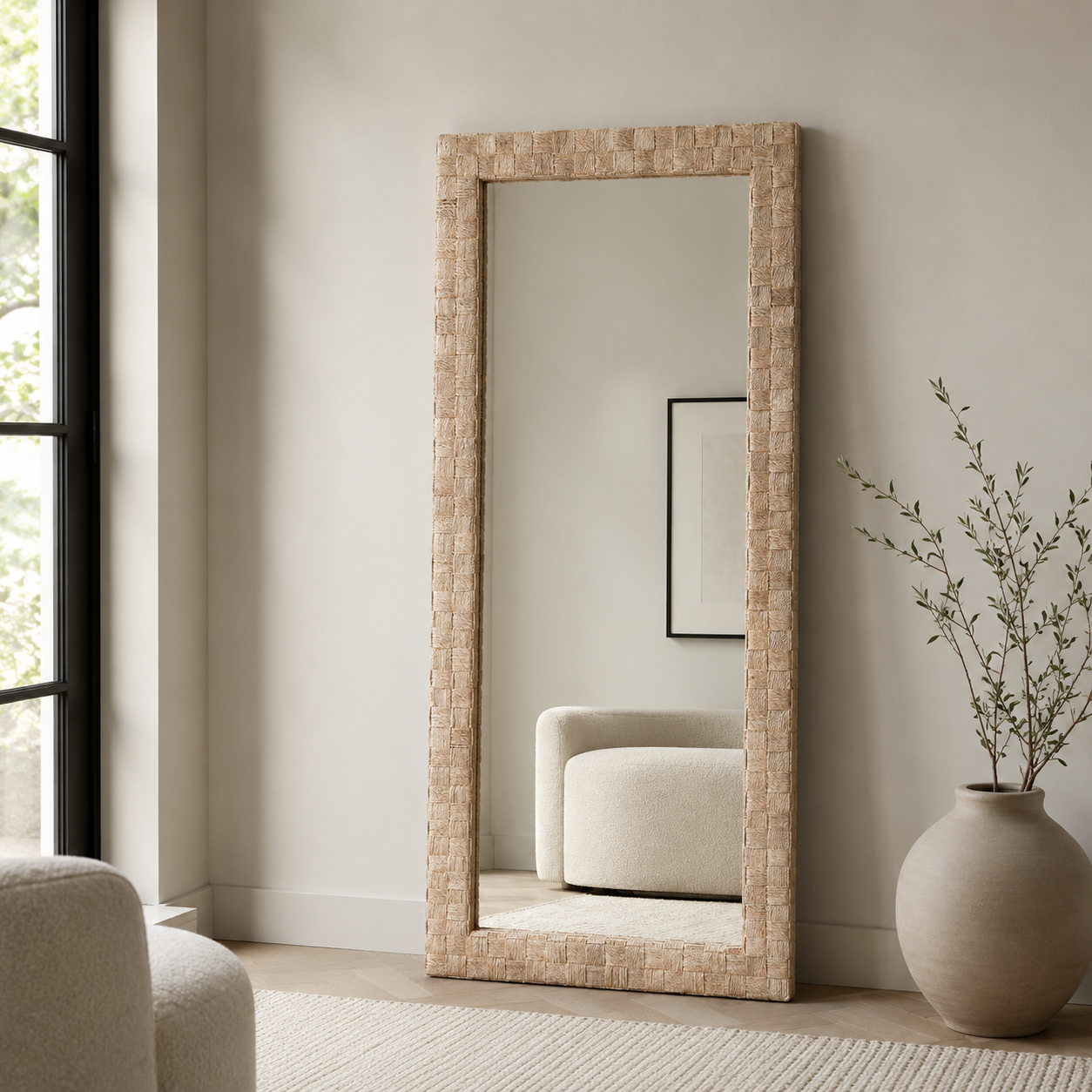 A tall floor mirror with a woven natural-fiber frame stands in a neutral, modern room next to a window, a beige armchair, and a large ceramic vase with dried branches.