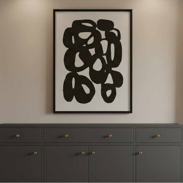 A dark gray sideboard cabinet in a styled room, decorated with a vase of pampas grass, books, a gold table lamp, and a large black and white abstract framed print on the wall above it.