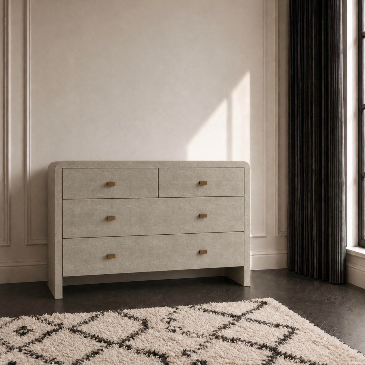 A light gray textured dresser with three drawers and small wooden knobs, positioned against a beige wall in a styled room with a dark rug and window curtains.
