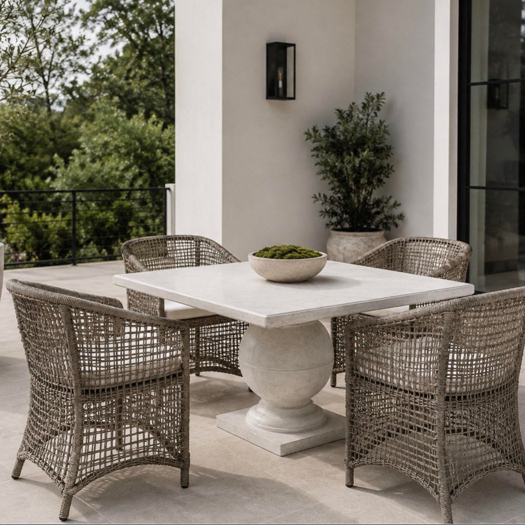 A 5-piece outdoor dining set featuring a square white stone table with a round pedestal base and four woven wicker chairs, set on a patio with a bowl of greenery on top.