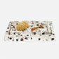 A rectangular hammered silver tray with a textured, organic surface pattern, styled with a natural tan loofah and a small orange-handled tool, on a clean white background.