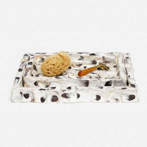 A rectangular hammered silver tray with a textured, organic surface pattern, styled with a natural tan loofah and a small orange-handled tool, on a clean white background.