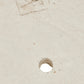 A close-up of a light beige textured surface with a small circular hole near the bottom center and a faint stamped square mark near the top left.