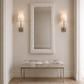 A tall rectangular wall mirror with a textured organic white frame hangs above a tufted bench with a light fabric seat and slim metallic legs in a softly lit entryway. Two matching wall sconces flank the mirror.
