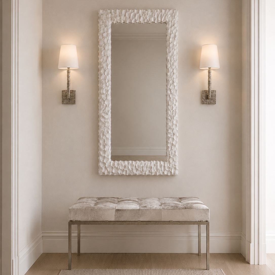 A tall rectangular wall mirror with a textured organic white frame hangs above a tufted bench with a light fabric seat and slim metallic legs in a softly lit entryway. Two matching wall sconces flank the mirror.