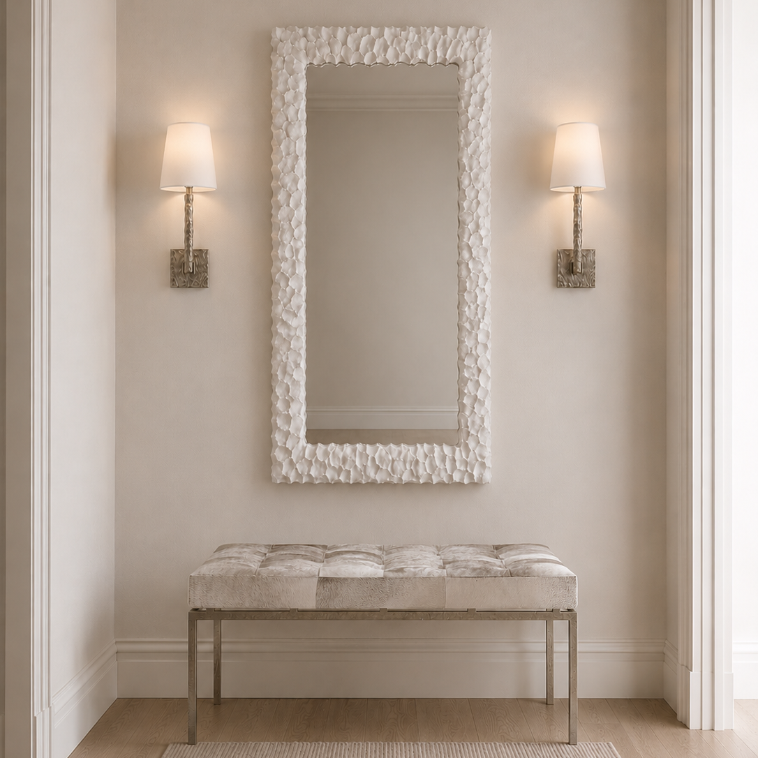 A tall rectangular wall mirror with a textured organic white frame hangs above a tufted bench with a light fabric seat and slim metallic legs in a softly lit entryway. Two matching wall sconces flank the mirror.