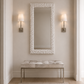 A tall rectangular wall mirror with a textured organic white frame hangs above a tufted bench with a light fabric seat and slim metallic legs in a softly lit entryway. Two matching wall sconces flank the mirror.