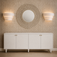 A white sideboard cabinet with four doors and small brass knobs, set against a textured beige wall. Above it is a large circular mirror with a beaded frame, flanked by two lit wall sconces.