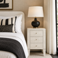 A modern bedroom scene featuring a light wood three-drawer nightstand beside a bed with white linens and a black throw blanket. The nightstand holds a black ceramic table lamp with a white drum shade and a gold base.