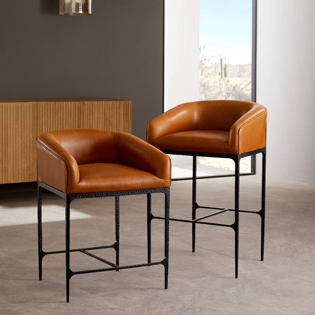 Two modern bar stools with warm brown leather seats and black metal frames, shown in a styled interior setting with a window and sideboard.