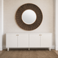 A large round wall mirror with a dark brown sunburst wood frame hangs on a textured beige wall above a white credenza with small gold knobs.