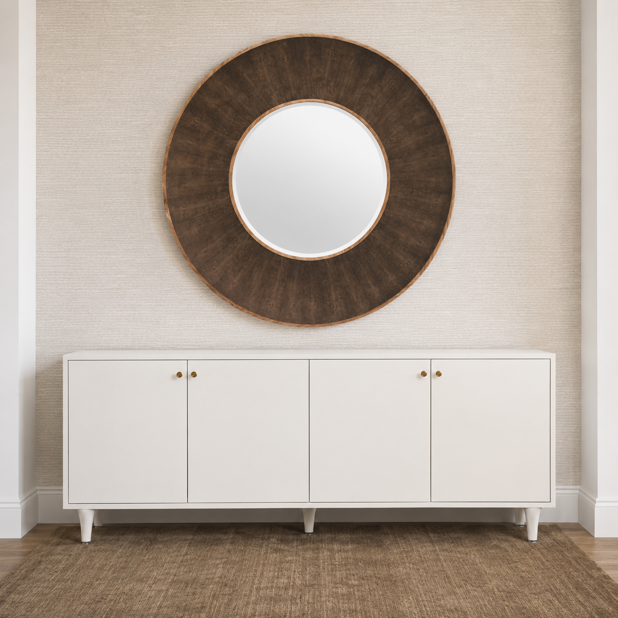 A large round wall mirror with a dark brown sunburst wood frame hangs on a textured beige wall above a white credenza with small gold knobs.
