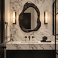Luxurious modern bathroom featuring a sculptural asymmetrical dark wood mirror, illuminated alabaster wall sconces, dramatic white marble walls and countertop, matte black vanity cabinetry, and dark bronze fixtures with refined black accents.