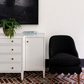 A styled interior corner featuring a white wooden cabinet with brass knobs, a black upholstered accent chair, a potted fern, and a large black framed wall art piece against a white wall.