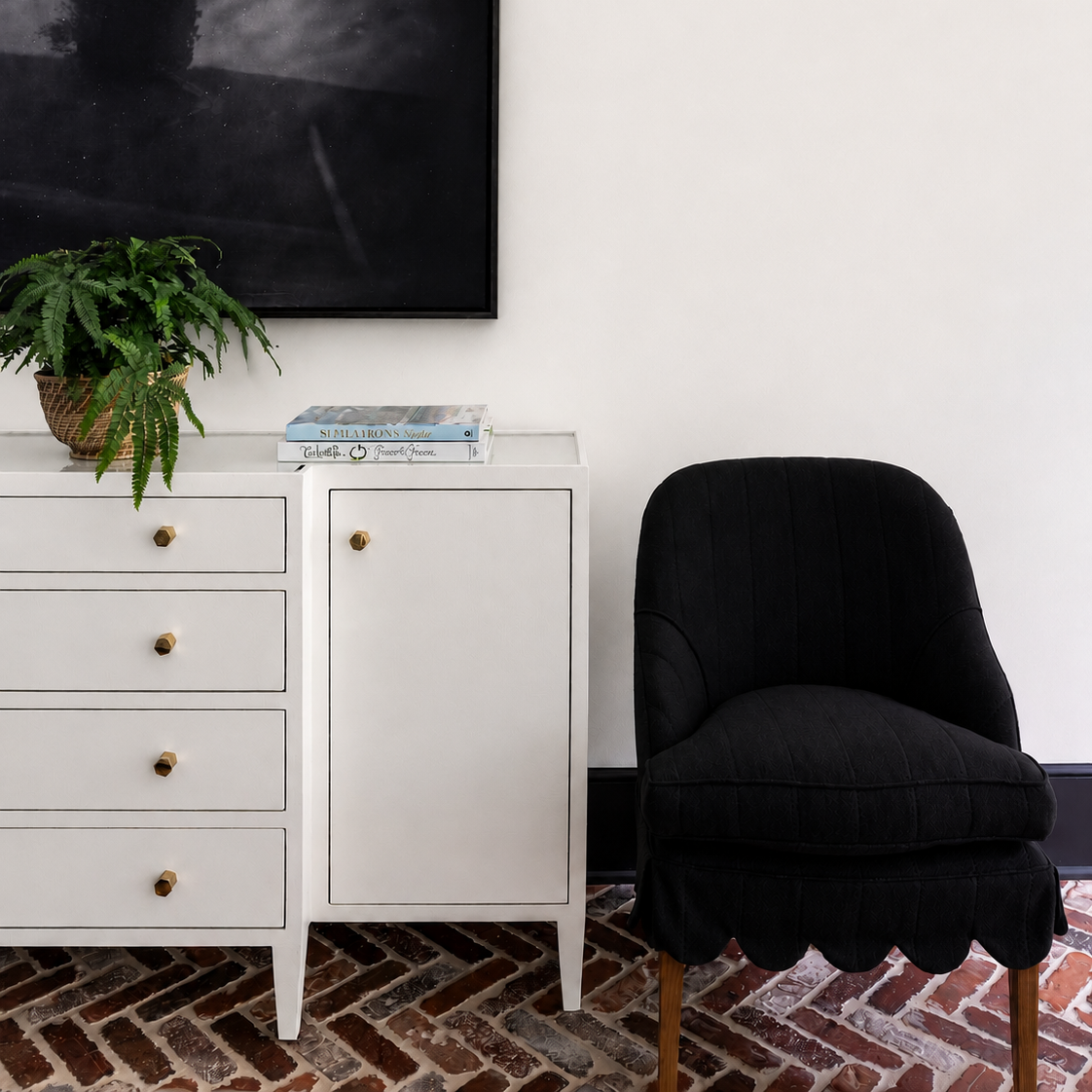 A styled interior corner featuring a white wooden cabinet with brass knobs, a black upholstered accent chair, a potted fern, and a large black framed wall art piece against a white wall.