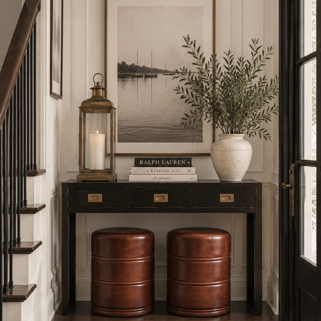 A styled entryway console table setup featuring a black wooden console, two brown leather barrel stools, a white ceramic vase with greenery, a brass lantern candle, and stacked books including a visible Ralph Lauren book.