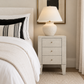 A modern bedside table in a light textured stone or concrete finish, styled in a bedroom next to a bed with white linens and a black-and-white striped pillow. The table features three drawers with small brass knobs and stands on tapered legs, topped with a matching textured table lamp with a white shade.