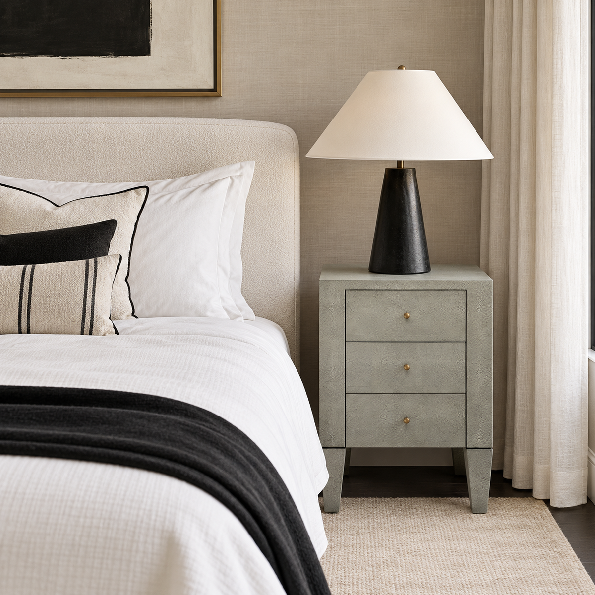 A modern bedroom scene featuring a light gray three-drawer nightstand with gold-tone knobs, topped with a black table lamp that has a white drum shade and a tapered black base. The nightstand sits beside a bed dressed in white textured bedding with a black throw blanket and decorative pillows, set against a warm beige wall with a curtain on the right.