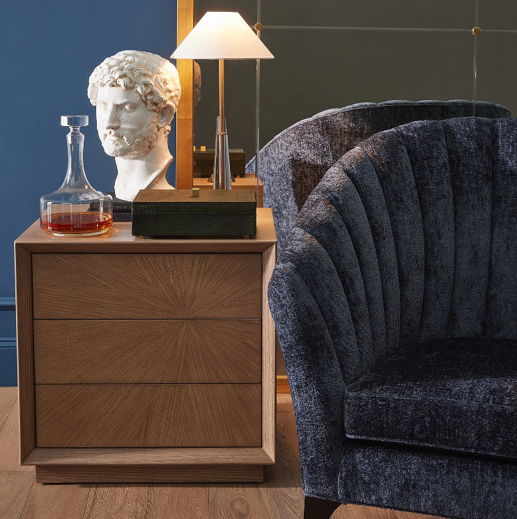 Wooden nightstand with decorative items next to a blue armchair against a dark wall.