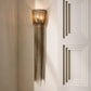 A modern wall sconce with a cylindrical woven shade and long hanging metallic fringe, mounted on a cream wall next to a white paneled door.