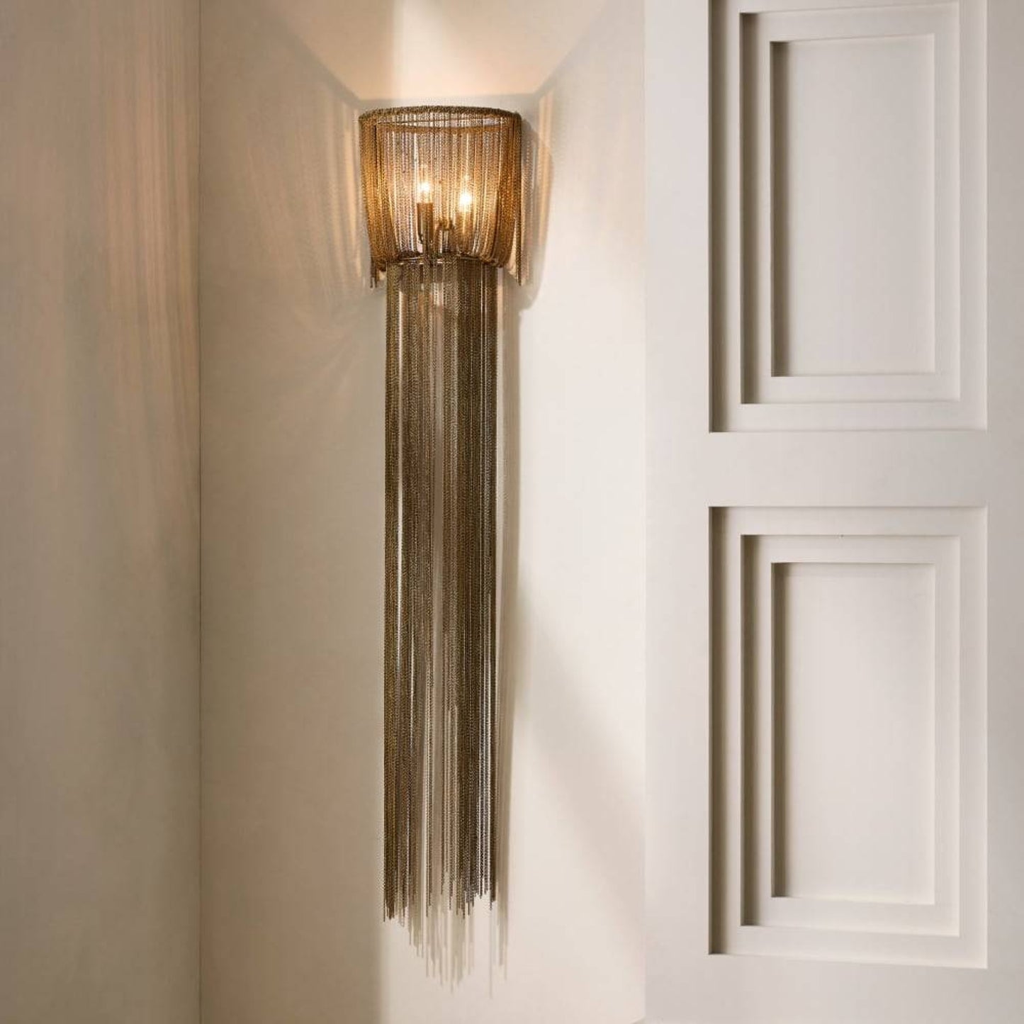 A modern wall sconce with a cylindrical woven shade and long hanging metallic fringe, mounted on a cream wall next to a white paneled door.