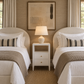 A symmetrical twin bedroom featuring two neatly made beds with white linens and taupe throws, flanking a white three-drawer nightstand with a lit ceramic table lamp. The room has a textured beige wall, a large abstract framed print, and cream drapes.