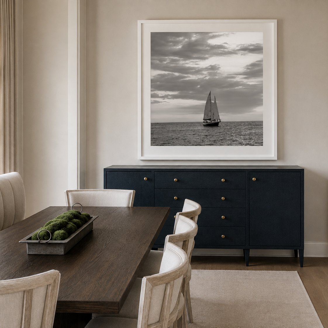 A large framed black and white art print of a sailboat on the water, displayed on a beige wall above a dark navy blue sideboard in a dining room setting.