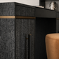 Close-up of a dark wood desk or cabinet with a textured black finish, a brass horizontal accent strip, and a vertical black handle, shown in an interior setting with part of a tan upholstered stool visible.