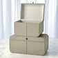 Two beige leather storage boxes stacked on a light surface with white curtains in the background