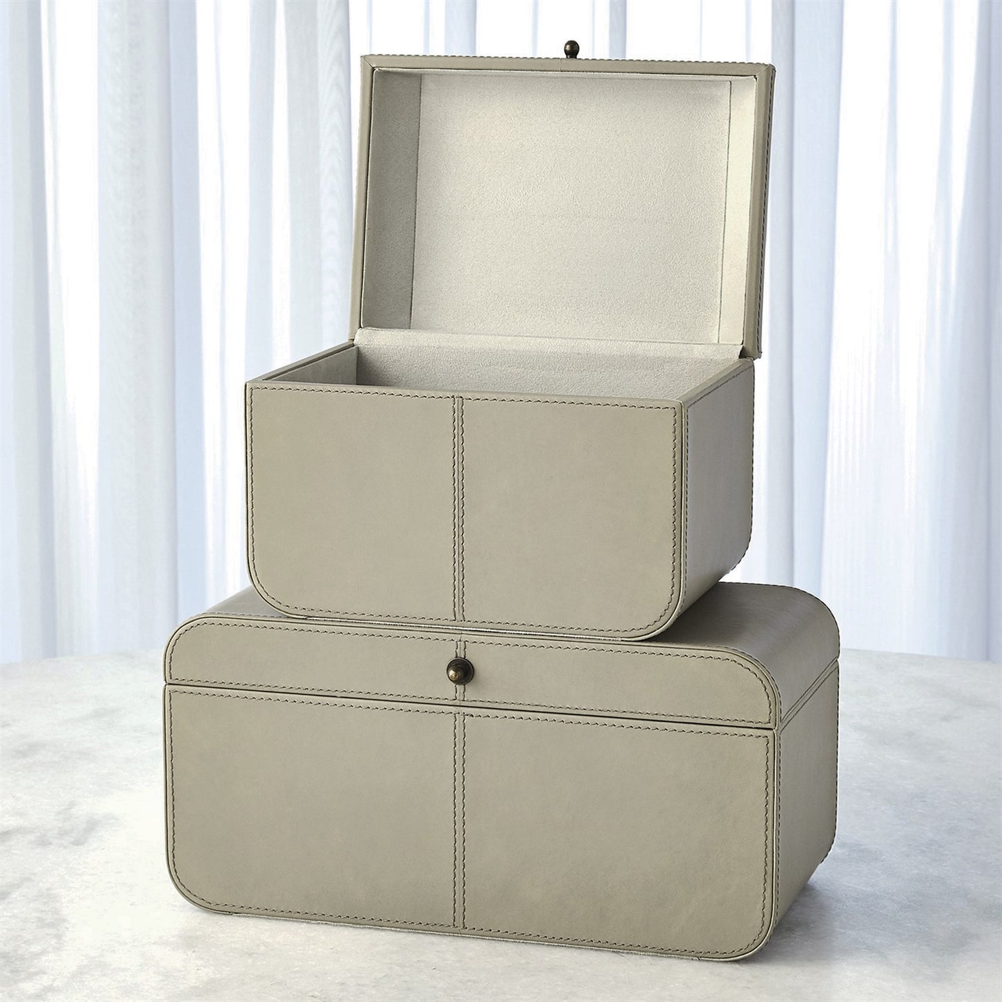 Two beige leather storage boxes stacked on a light surface with white curtains in the background