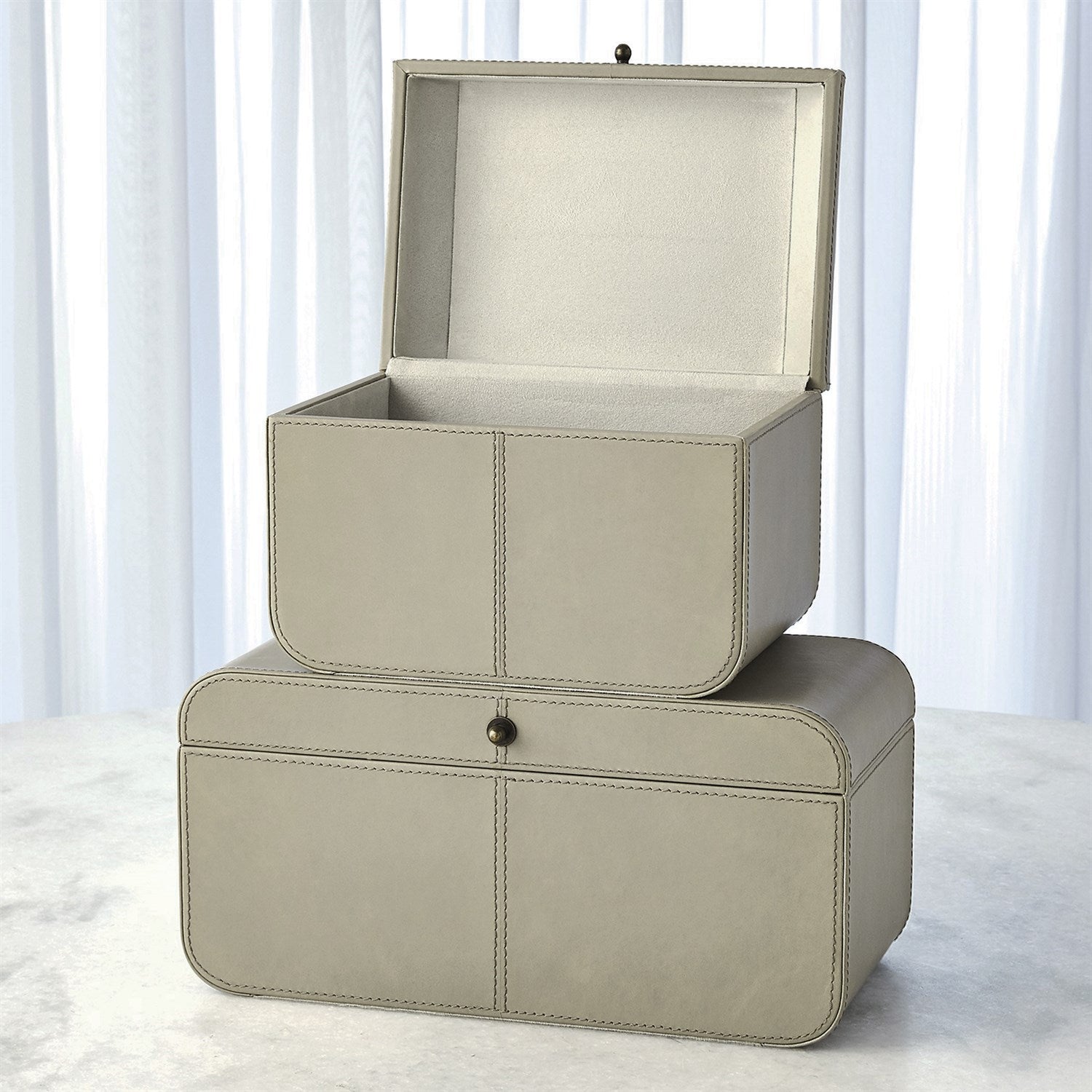 Two beige leather storage boxes stacked on a light surface with white curtains in the background