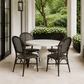 A round white dining table with four dark woven wicker chairs, set on a light stone patio under a covered porch with green garden foliage in the background.