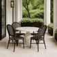 A round white dining table with four dark woven wicker chairs, set on a light stone patio under a covered porch with green garden foliage in the background.