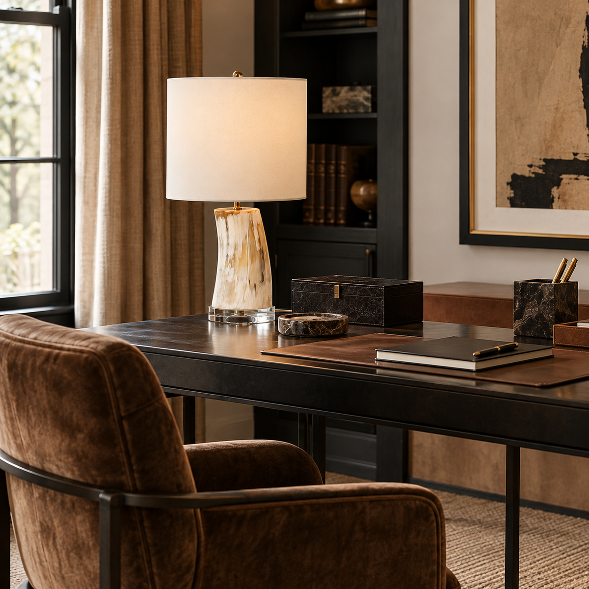 Sophisticated modern home office featuring a sculptural cream and amber marble table lamp with an ivory drum shade and gold accents, styled on a dark wood desk alongside black marble accessories, a leather desk pad, and a brown velvet chair. Black built-in shelving, abstract artwork, and natural window light complete the elegant, masculine space.