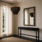 A styled interior room scene showing a black tiered chandelier hanging in a window with sheer curtains, a black console table beneath a patterned rectangular mirror, and the chandelier reflected in the mirror.