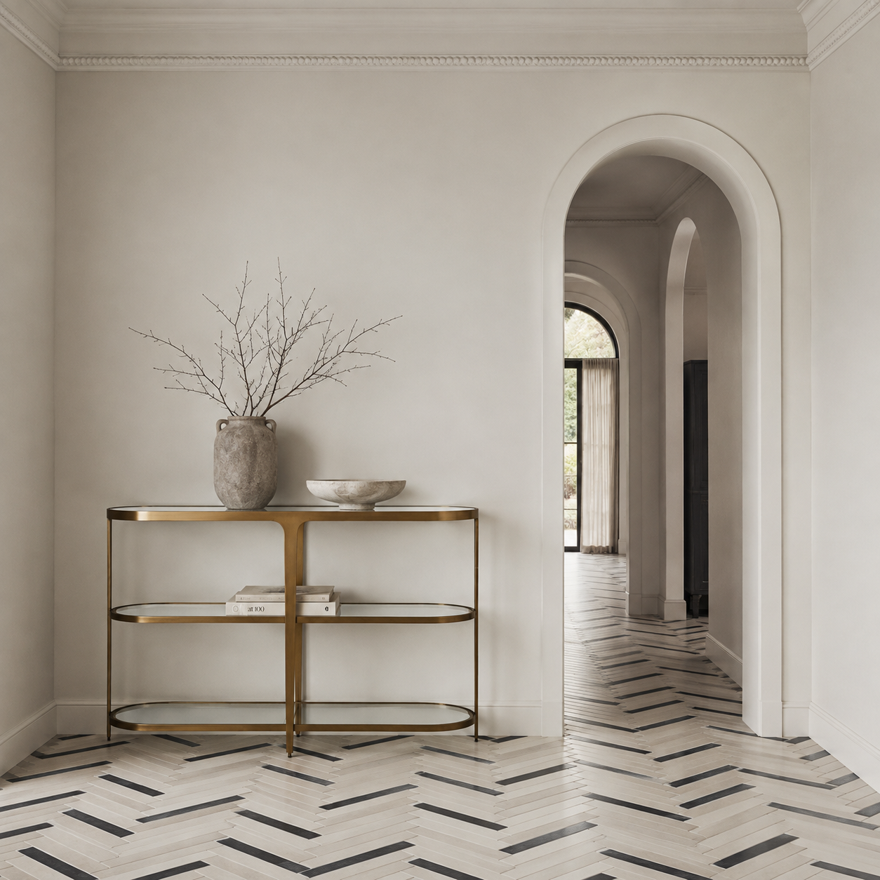 A modern gold-framed console table with glass shelves in a minimalist hallway featuring a herringbone wood floor and an arched doorway.