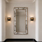 A tall rectangular wall mirror with a distressed white and gray marbled frame, mounted on a beige wall between two lit black sconces.