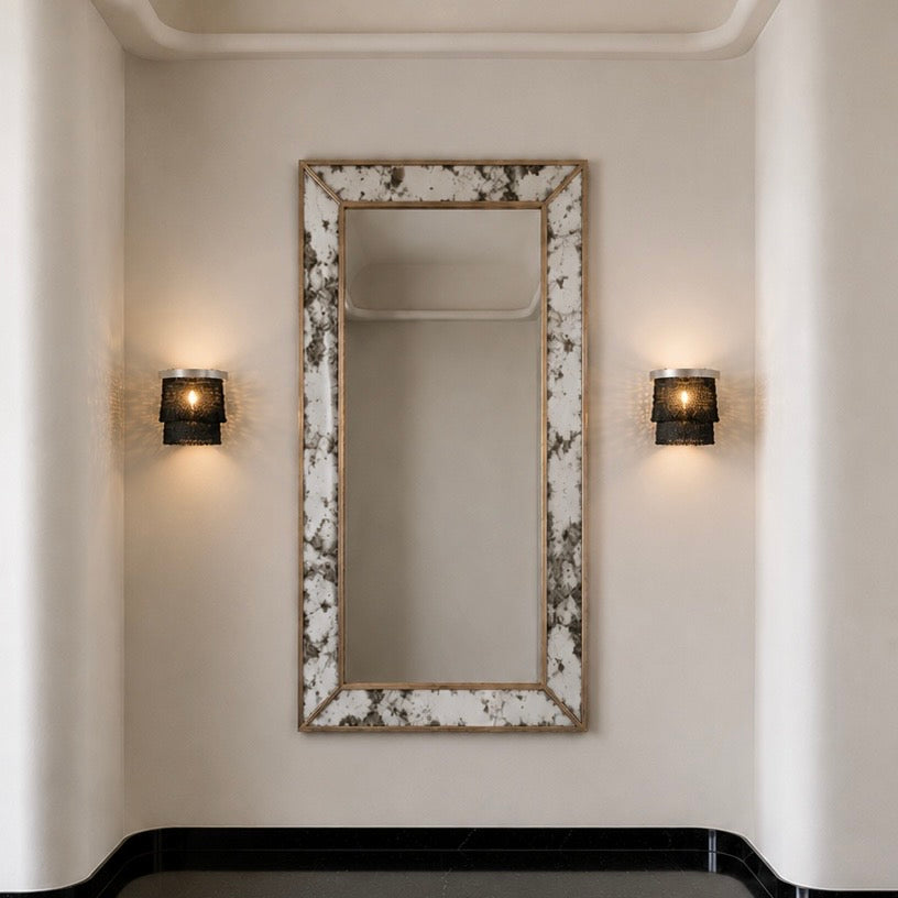 A tall rectangular wall mirror with a distressed white and gray marbled frame, mounted on a beige wall between two lit black sconces.