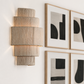 A modern tiered wall sconce with a textured beige linen fabric shade, mounted on a white wall next to framed abstract black and gray art.