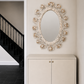 An ornate oval wall mirror with a sculptural floral frame in warm beige and cream tones, mounted above a matching cream cabinet in a hallway with dark tiled flooring and a black staircase visible in the background.