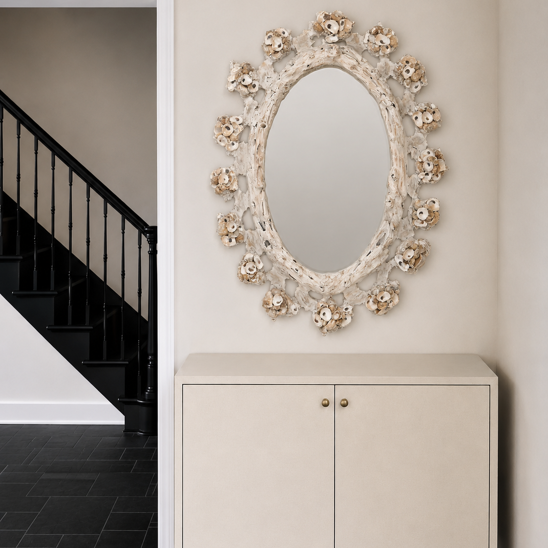 An ornate oval wall mirror with a sculptural floral frame in warm beige and cream tones, mounted above a matching cream cabinet in a hallway with dark tiled flooring and a black staircase visible in the background.