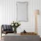 A rectangular wall mirror with a distressed silver frame hangs on a white paneled wall above a gray console table. The scene is styled with a brown leather chair, a vase of white flowers, a bamboo tray, and a tall floor lamp.