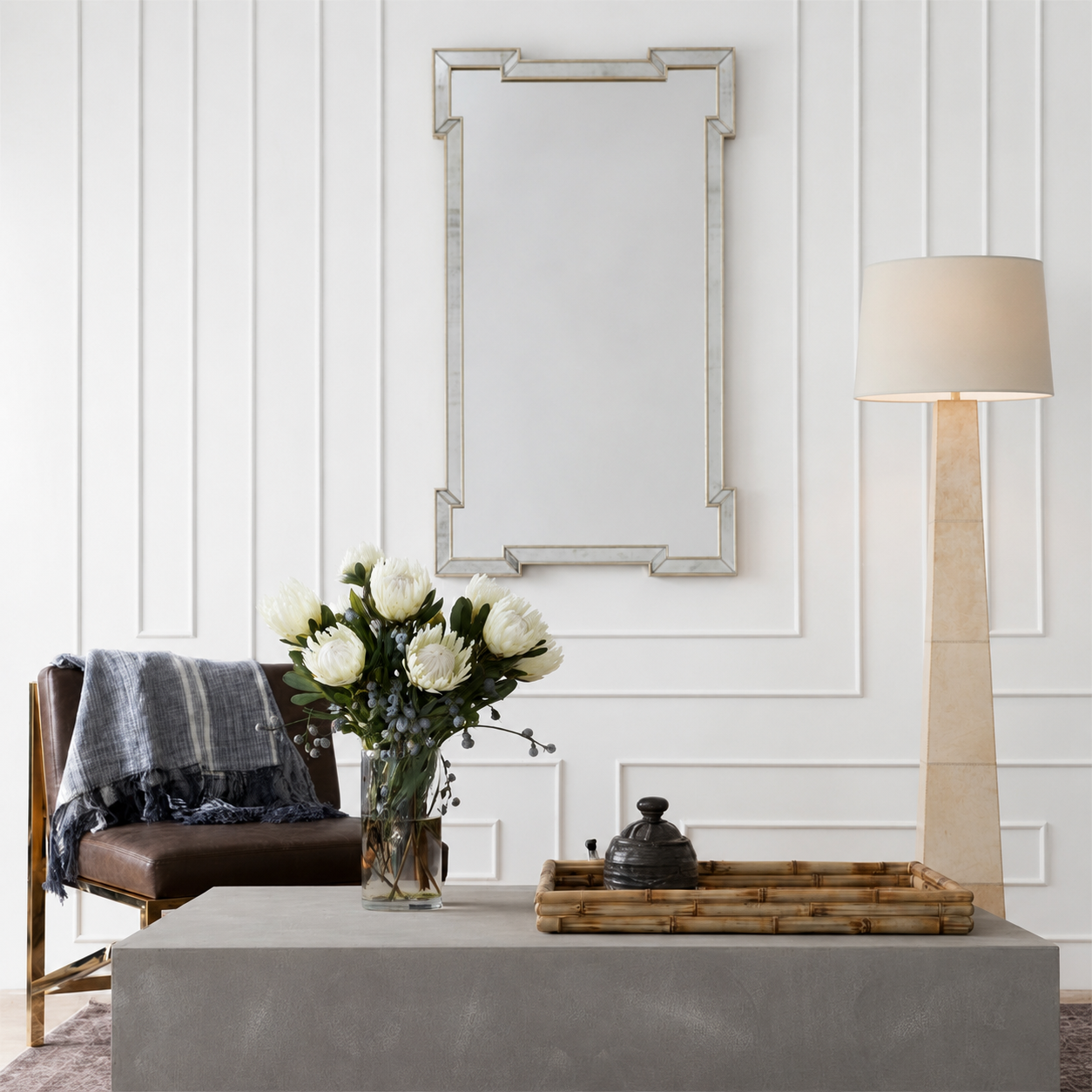 A rectangular wall mirror with a distressed silver frame hangs on a white paneled wall above a gray console table. The scene is styled with a brown leather chair, a vase of white flowers, a bamboo tray, and a tall floor lamp.