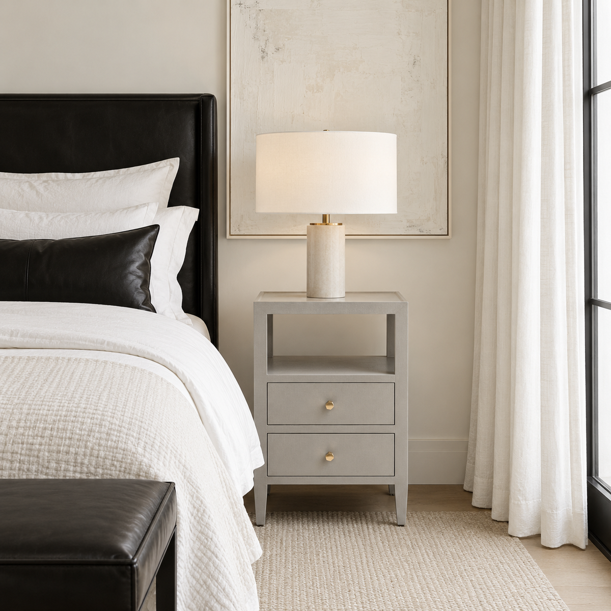 A modern bedroom scene featuring a light gray three-drawer nightstand with gold knobs, styled with a table lamp on top. The nightstand is positioned next to a bed with white bedding and a black leather headboard, set against a neutral beige wall with a large framed print and sheer white curtains.