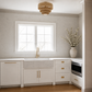 A bright, modern kitchen corner with white cabinetry, a white farmhouse sink, a gold faucet, a window, a round wood pendant light, and a large vase with branches on the counter.